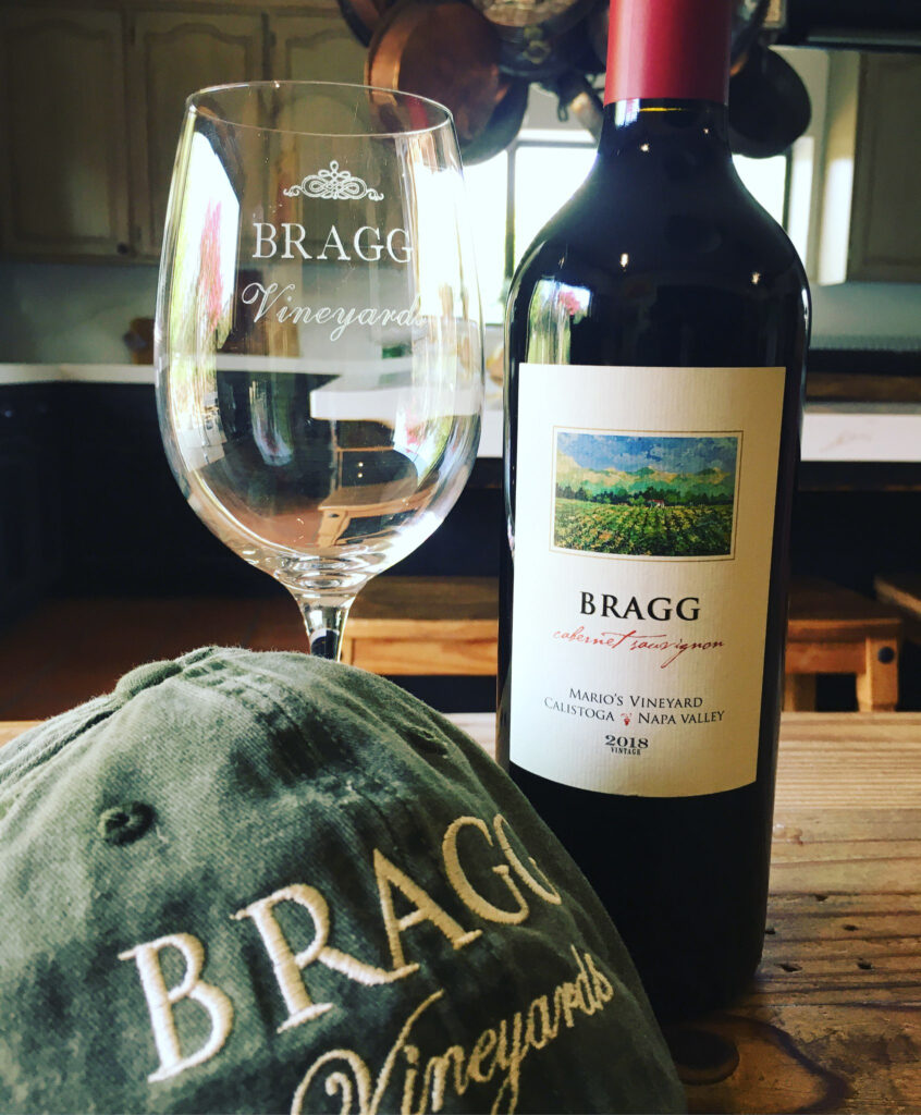 bottle of Bragg Vineyards wine on a wooden dining table with a wine glass and logo hat